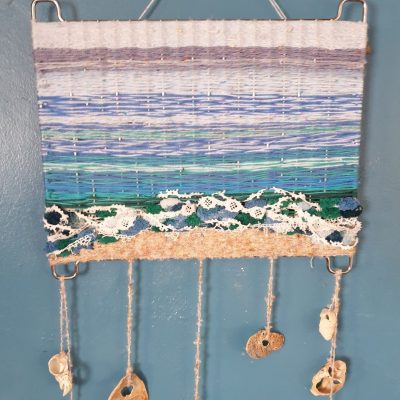 A seascape weaved into a metal grill, with portions of horizon, sea, foam, and beach. Attached shells dangle below.
