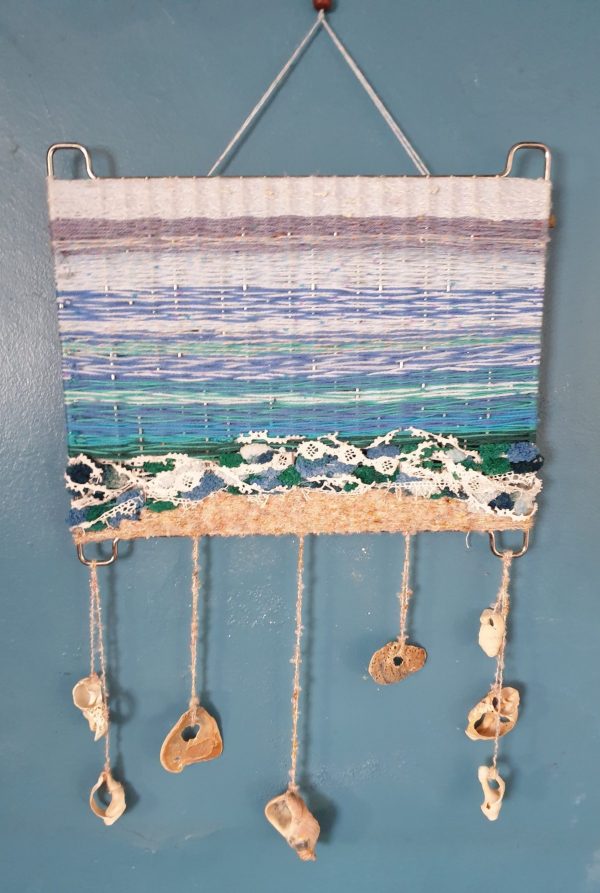 A seascape weaved into a metal grill, with portions of horizon, sea, foam, and beach. Attached shells dangle below.